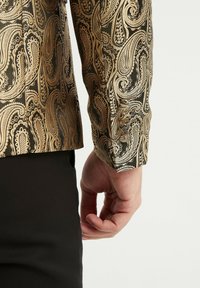 Chemise à motif paisley doré sur une base noire, présentant une texture brillante et des détails de poignet à boutons. Main positionnée près de la taille.