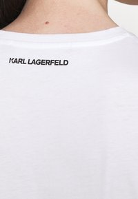 Biała bawełniana koszulka z okrągłym dekoltem. Z tyłu znajduje się czarny nadruk z logo "KARL LAGERFELD". Miękka tekstura, luźny fason.