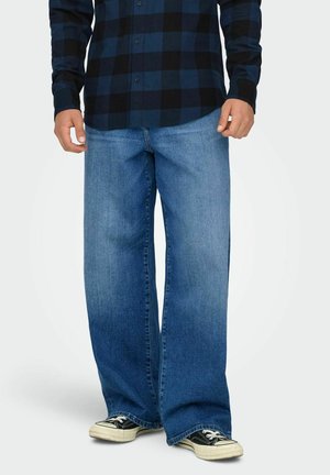 Persoană îmbrăcată cu blugi largi albaștri, cămașă în carouri albastre și negre și adidași negri cu alb, stând pe un fundal simplu.