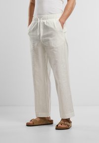 Ľahké, textúrované nohavice v drevno-bielej farbe s elastickým pásom a bočnými vreckami, v kombinácii s hnedými sandálmi s nastaviteľným remienkom.