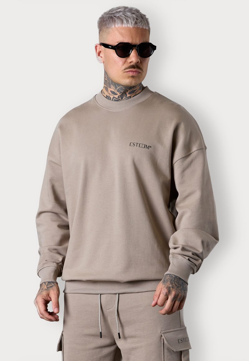 Sweatshirt beige en tissu doux avec un col rond et des poignets côtelés. Présente un petit logo "ESTEEM" sur la poitrine et une coupe décontractée.