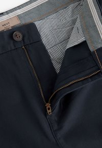 Pantaloni slim fit blu navy scuro parzialmente slacciati, che mostrano la fascia interna a righe e un bottone scuro allacciato in alto.