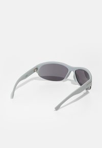 Gafas de sol deportivas con un marco de plástico gris claro, lentes gris oscuro, diseño curvado y patillas flexibles. Presentan un pequeño acento metálico.