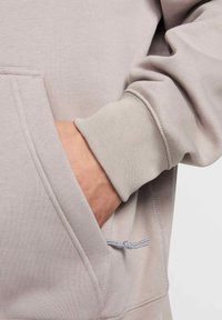 Hoodie gris clair en tissu texturé, doté d'une poche avant et d'un cordon de serrage. Les poignets sont côtelés, ajoutant élasticité et structure.