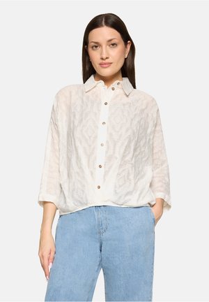 Femme aux cheveux lisses et foncés portant un chemisier blanc transparent à boutons et un jean taille haute bleu clair, posant avec une main dans la poche.