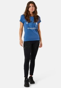 Blå bomullströja med vita grafiska detaljer, parat med svarta tajta byxor med fickor. Modellen bär svarta sneakers.