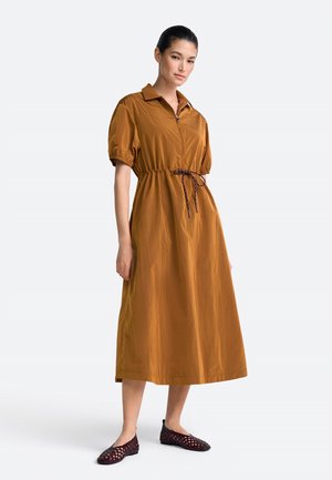 Nő rozsdabarna midi ruhában, rövid pufók ujjakkal, gallérral, elülső cipzárral és megkötős derékrésszel, sötét szövött lapos cipővel párosítva.