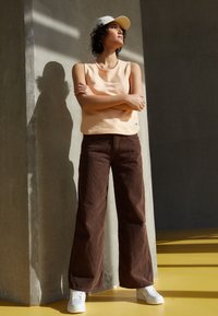 Meruňkový top bez rukávů, hnědé kordové kalhoty širokého střihu a bílé tenisky; outfit doplňuje neutrální kšiltovka a čisté linie.