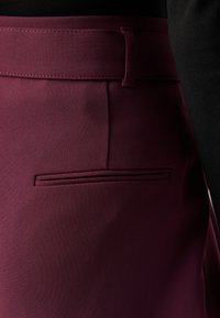 Bordeaux bukser med glatt stoff, med en flat midje, en dekorativ lomme og subtil søm. Detaljert tekstur synlig.