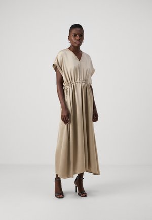 Robe maxi beige en tissu doux, avec un décolleté en V, des manches courtes et une taille cintrée, accompagnée de sandales à talons noires.