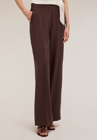 Un pantalon marron long et ample en tissu texturé avec des poches latérales et une taille plate, associé à des sandales à lanières.