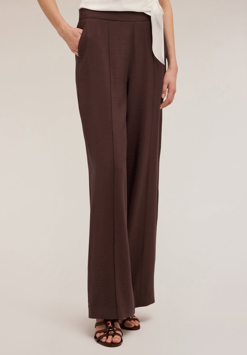 Un pantalon marron long et ample en tissu texturé avec des poches latérales et une taille plate, associé à des sandales à lanières.