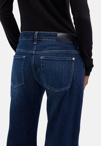 Ciemnoniebieskie jeansy z denimu o wysokim stanie, wyposażone w teksturowaną czarną skórzaną łatę z tyłu oraz podwójne tylne kieszenie z kontrastowym przeszyciem.