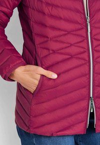 Main dans la poche d'une veste matelassée bordeaux avec des coutures diagonales et une fermeture éclair frontale en argent, portée avec un jean bleu.