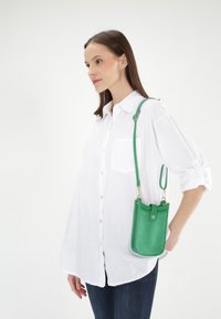 Grænt læder crossbody taske med en struktureret form, guldbeslag og en sikker lukning. Båret med en hvid skjorte med knapper.