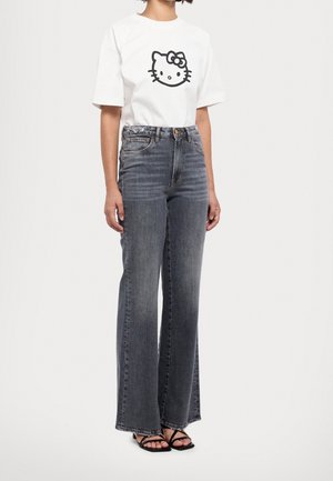 Woman wearing a white T-shirt with a black Hello Kitty face and high-waisted faded gray wide-leg jeans with black strappy sandals.