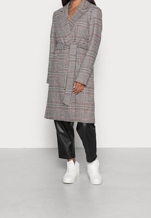 Femme portant un manteau à carreaux ceinturé, un pantalon en similicuir noir et des baskets montantes blanches, debout devant un fond blanc uni.