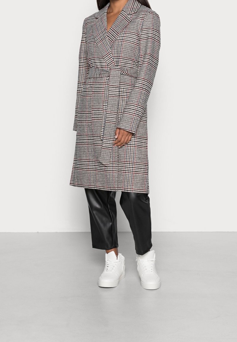 Femme portant un manteau à carreaux ceinturé, un pantalon en similicuir noir et des baskets montantes blanches, debout devant un fond blanc uni.