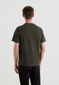 Camiseta de rayas verdes con cuello redondo, mangas cortas y una textura suave. Las rayas son de color verde oscuro y negro. Pantalones negros visibles.