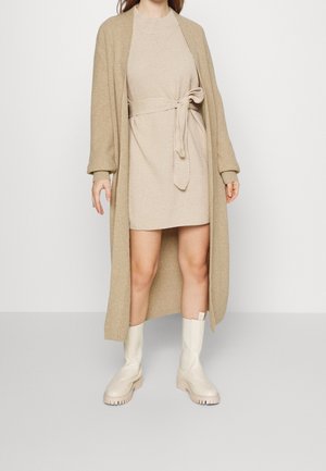 Femme portant une robe en maille beige avec ceinture, un long cardigan beige et des bottes épaisses mi-mollet de couleur blanc cassé, debout contre un mur uni.
