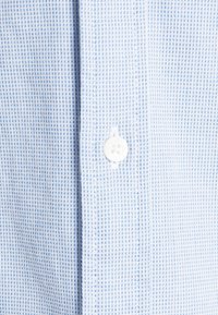 Lichtblauw geruit overhemdstof met een witte knoop in het midden. De textuur vertoont kleine, consistente patronen door het hele oppervlak.