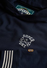 Felpa blu navy in cotone con logo frontale bianco, che presenta un anno e assi incrociati; include dettagli di accenti a strisce sui polsini e etichetta.