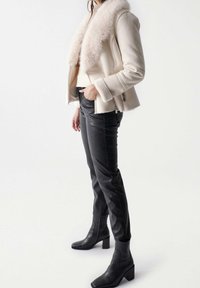 Weiße Jacke mit Kunstfellkragen und taillierter Passform, kombiniert mit schwarzen Lederhosen und Ankle Boots. Die Jacke verfügt über Reißverschlusstaschen.