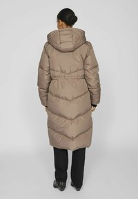 Manteau rembourré long et brun avec une capuche et une taille élastique. Présente un matelassage en chevron et un design jusqu'aux genoux. Porté avec un pantalon noir.