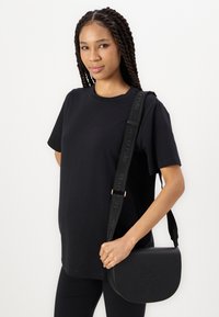 Čierna kožená crossbody taška zaobleného tvaru, s embosovaným logom a nastaviteľným popruhom so značkou. Nosí sa s jednoduchým čiernym tričkom.