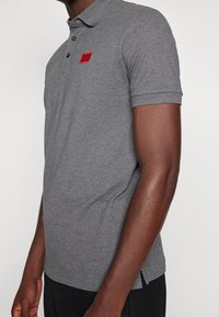 Camisa polo de manga corta gris con cuello acanalado, dos botones y un parche de logo rojo en el lado izquierdo del pecho. Aberturas laterales en el dobladillo.