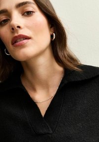 Czarny dzianinowy sweter z dekoltem w szpic, srebrny łańcuszek i kolczyki w kształcie kół. Gładka tekstura i swobodny fason, częściowo przycięta twarz.