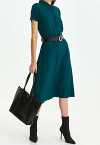Teal kortärmad klänning med knappar framtill, åtdragen i midjan med ett svart bälte med ett runt guldspänne. Kombineras med svarta ankelskor och en tote-väska.