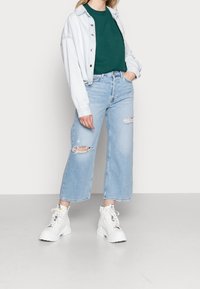Jeans a gamba larga blu chiara con dettagli strappati e orli sfrangiati, abbinati a una maglietta verde e a una giacca di denim chiaro. Sneakers bianche chunky.