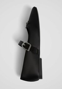 Chaussure plate en satin noir avec un bout pointu et une sangle à boucle en argent, photographiée sur un fond blanc uni.