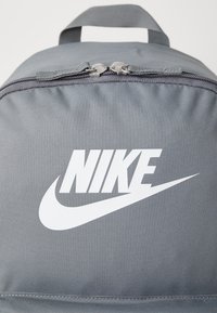 Mochila cinza em nylon com fechos duplos, apresentando um logo e swoosh da Nike em branco destacados no painel frontal. Textura suave, design compacto.