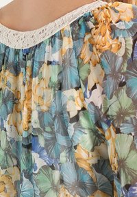 Blomstret bluse med en gennemsigtig, letvægtsstof med blå, grønne, gule og hvide blomstermønstre. Kantet med en sart blondeudskæring.