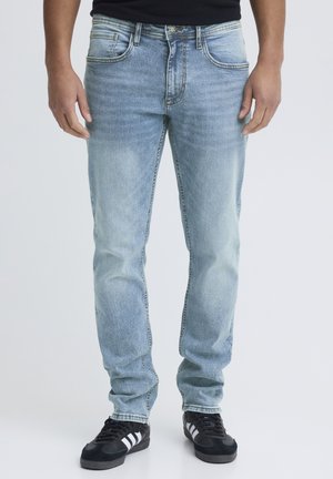 Mann steht und trägt hellblaue Slim-Fit-Jeans sowie schwarze Sneakers mit weißen Streifen, vor einem schlichten hellen Hintergrund.