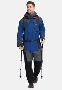 Uomo che fa escursionismo con giacca e pantaloni blu e grigi, con uno zaino e tenendo in mano bastoncini da trekking, cammina avanti sorridendo.