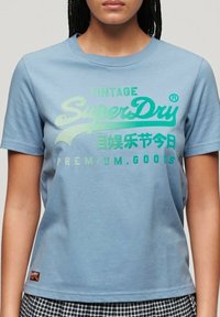 Ljusblå bomulls-t-shirt med rund halsringning. Har grön text: "VINTAGE SuperDry" och "PREMIUM GOODS" med logotyp. Kort ärm.