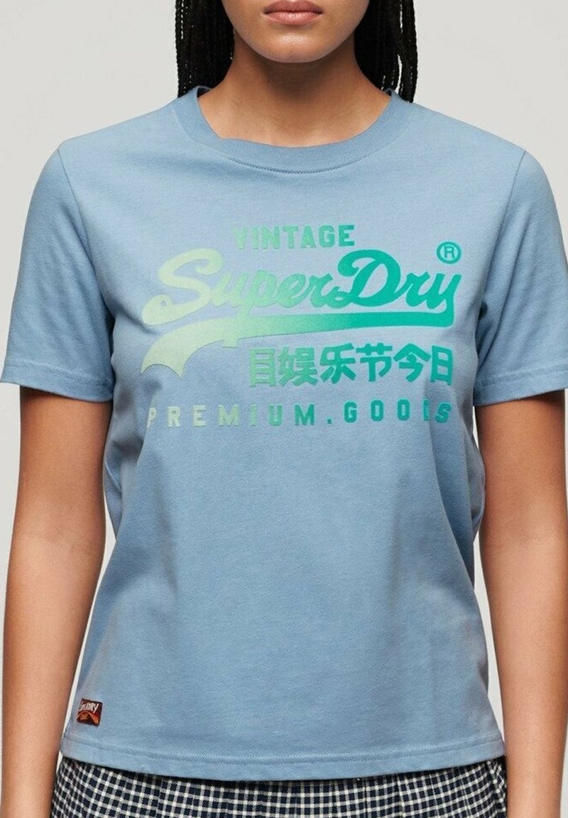 Ljusblå bomulls-t-shirt med rund halsringning. Har grön text: "VINTAGE SuperDry" och "PREMIUM GOODS" med logotyp. Kort ärm.