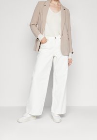 Světle béžový blejzr přes měkký krémový svetr, doplněný širokými bílými kalhotami a bílými teniskami. Hladké textury, precizní střih.