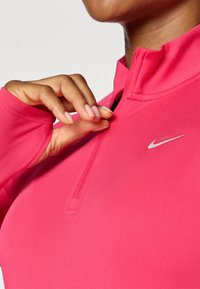 Fuksija atletska majica s četvrtim zatvaračem, mekog materijala i malim bijelim Nike logom na prsima. Kratki prikaz ruke na zatvaraču.