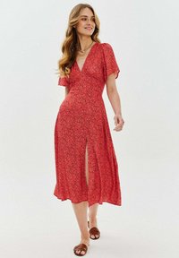 Červené květinové šaty s krátkými volánkovými rukávy, hlubokým výstřihem do V a předním rozparkem. Lehká látka s volně splývavým designem. Nosené hnědé sandály.