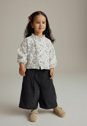 Cămașă albă cu flori, pantaloni negri lejeri și pantofi slip-on bej. Caracteristici includ guler ondulat și material patternat.