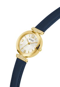 Montre dorée avec un cadran rond blanc, bracelet en cuir bleu marine et accents en cristal brillant sur les cornes. Présente de grands index et aiguilles.