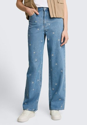 Persona che indossa jeans azzurri a gamba larga con piccoli ricami floreali bianchi, sneakers bianche, giacca beige senza maniche e top bianco.