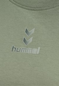 Gros plan d’un tissu gris-vert avec le nom de la marque "hummel" et deux flèches en chevron imprimées au-dessus.
