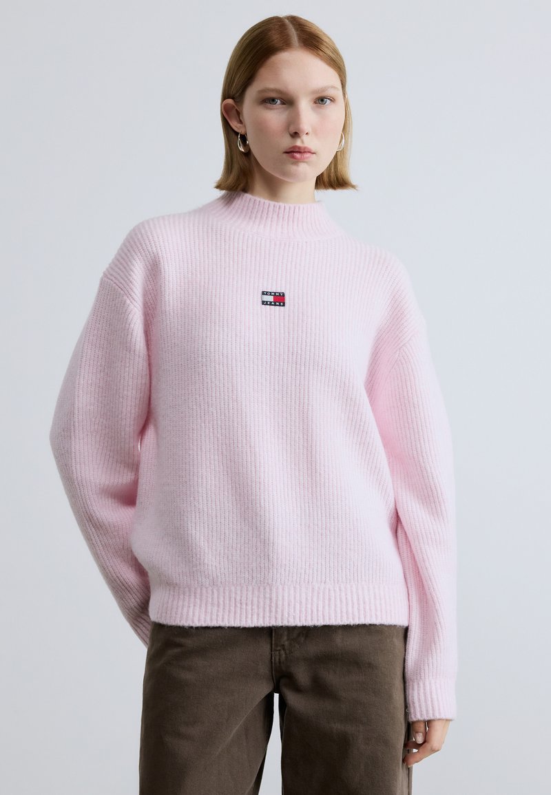 Pulover roz cu dungi, guler înalt, cu o mică emblemă pe piept și textură tricotată. Asortat cu pantaloni maro.