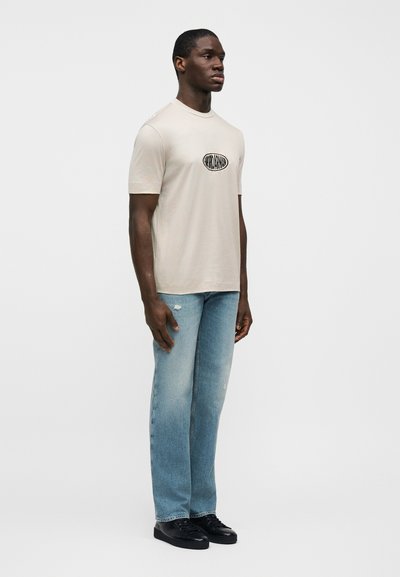 Μπεζ βαμβακερό μπλουζάκι με στρογγυλή λαιμόκοψη και μαύρο οβάλ λογότυπο, συνδυασμένο με ανοιχτόχρωμο μπλε τζιν παντελόνι και μαύρα αθλητικά παπούτσια, στέκεται μπροστά από ουδέτερο φόντο.