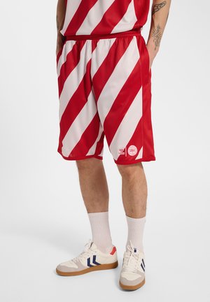 Person iført røde og hvide diagonalt stripede sportsshorts, hvide sokker og hvide sneakers med blå og røde accenter, står med hænderne i lommerne.